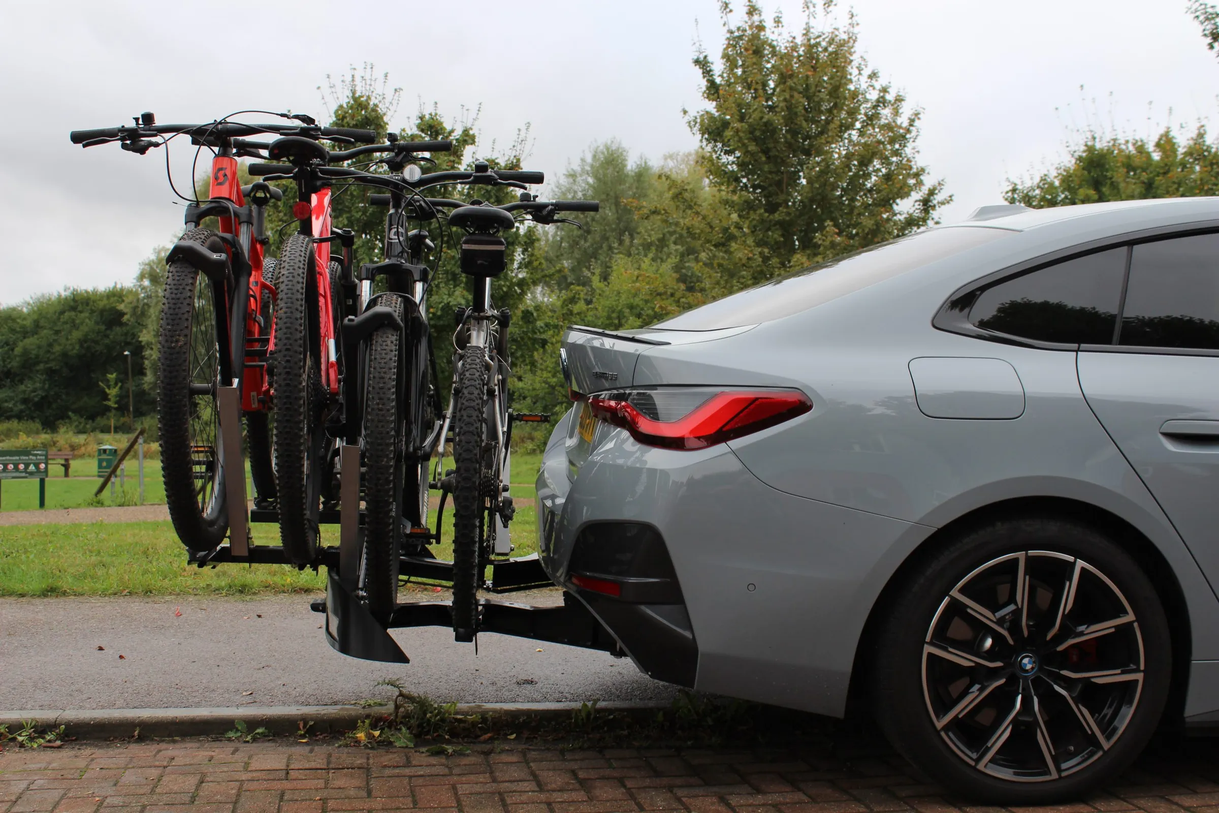 A silver BMW i4 parked at a kerb beside a park, with the Auxtail rack extended behind it carrying 4 mountain bikes upright and clear of the bodywork.