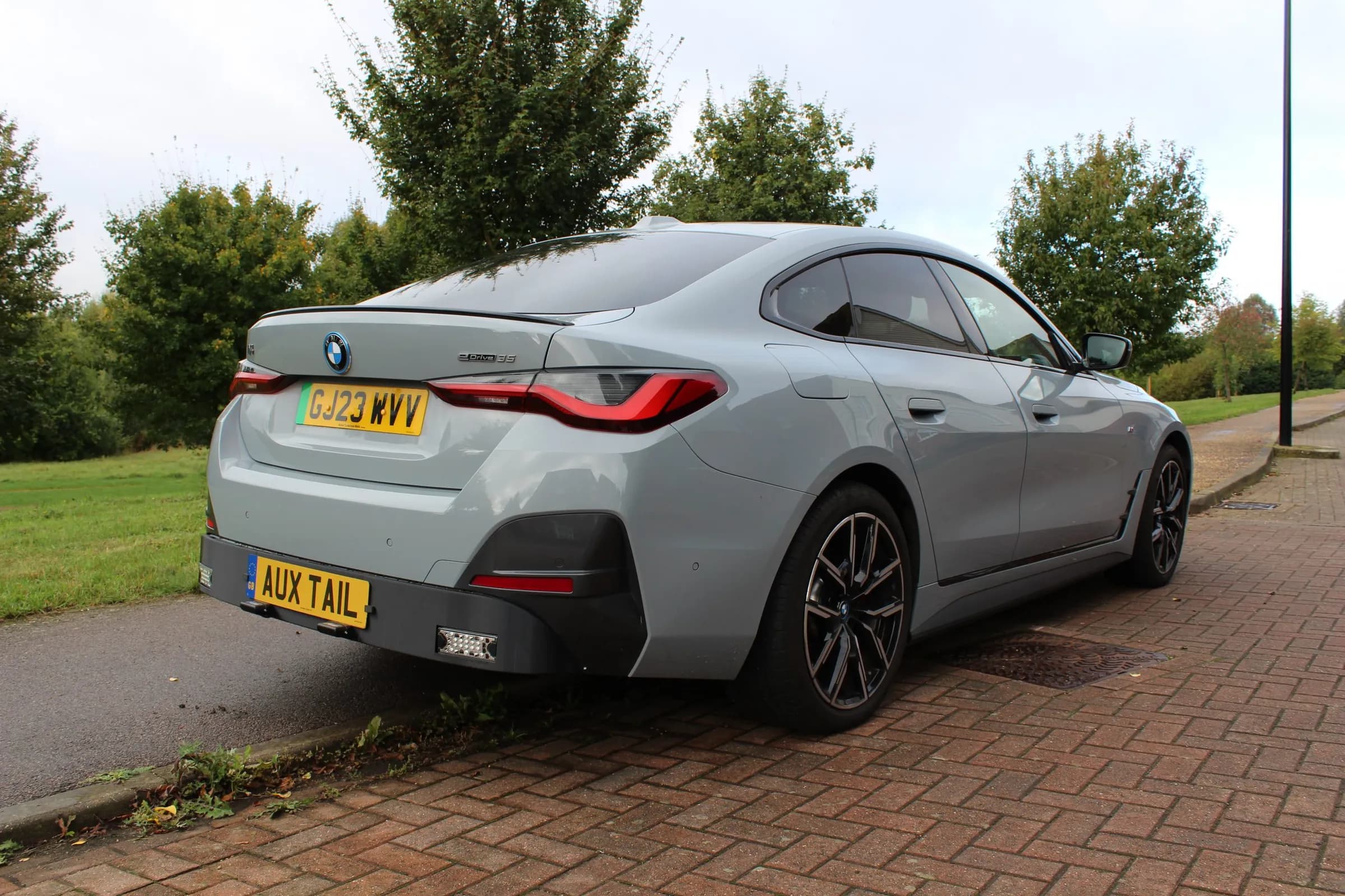 The same silver BMW i4 from behind with no bikes on it. The Auxtail rack is folded flat across the rear bumper, carrying the number plate and its own lights, and reads as part of the car.
