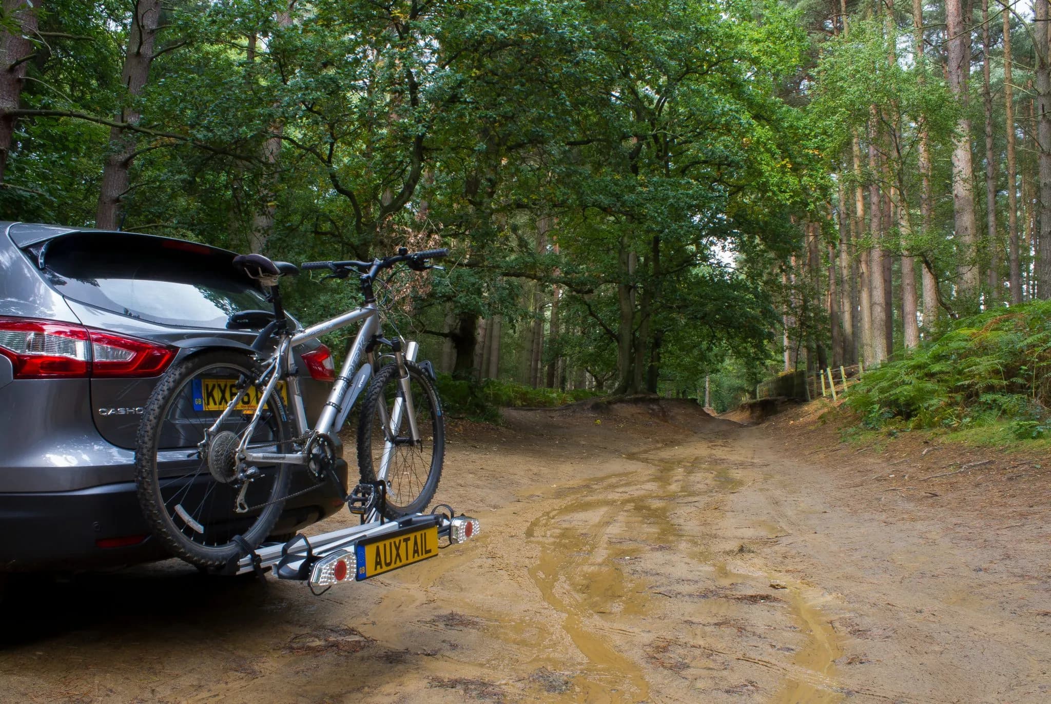 A grey Nissan Qashqai parked at the edge of a sandy forest track, carrying 1 mountain bike on the Auxtail rack, with the rack's own lighting board and AUXTAIL plate below the bike.