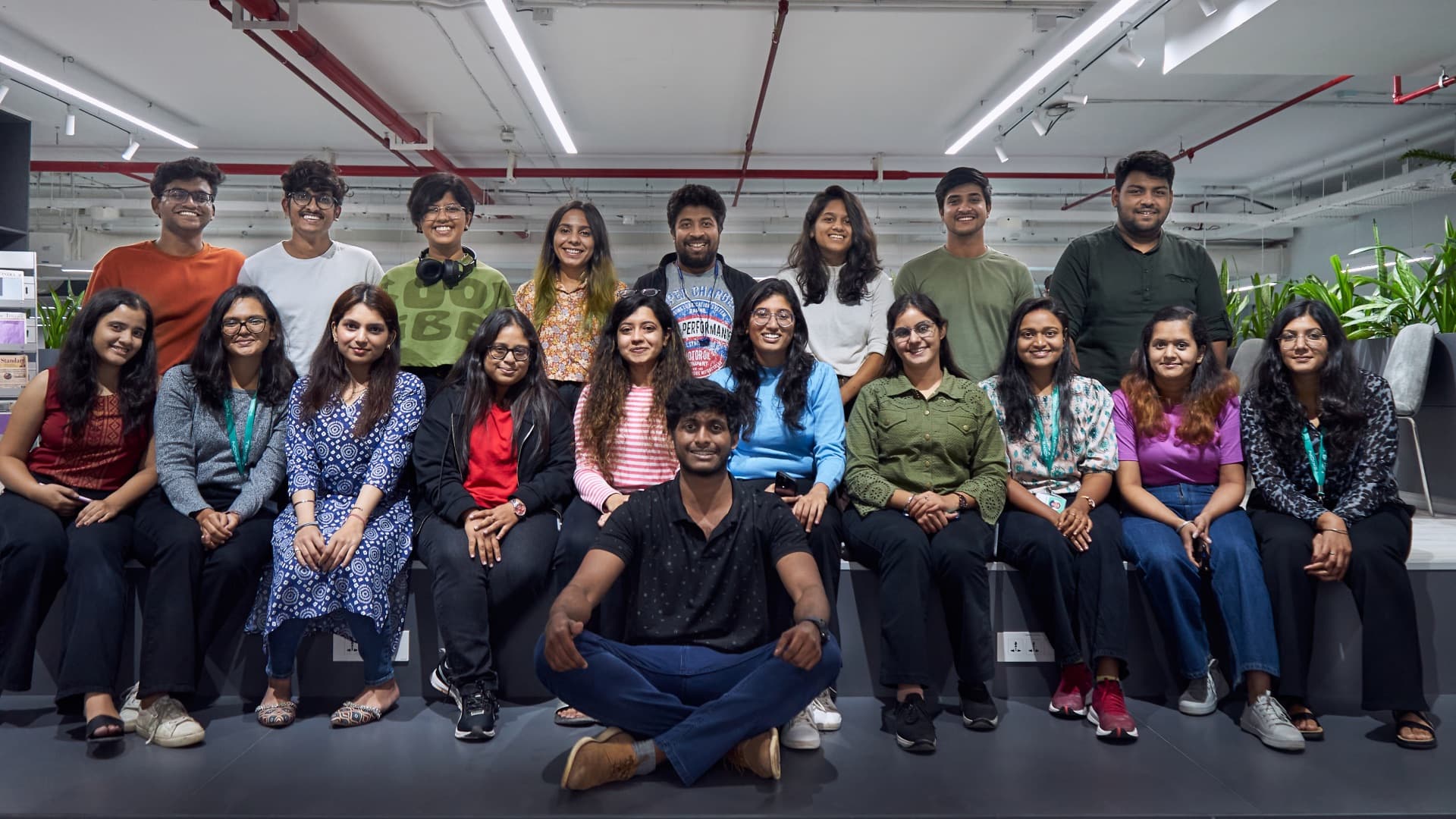 A group photographed indoors, seated and standing along a low step, with one person sitting cross legged at the front.